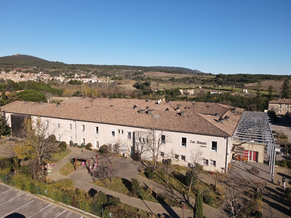 Vue aérienne de l'Ehpad Los Ainats avec ses toits en tuiles, son jardin arboré et ses espaces extérieurs aménagés, entouré par le paysage vallonné de l'Aude.