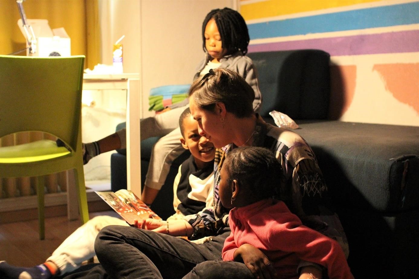 Une femme lit un livre à deux jeunes enfants assis près d'elle, dans une pièce chaleureusement éclairée, tandis qu'un autre enfant observe en arrière-plan.